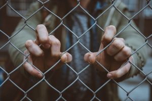 A person stands behind a wire fence with their hands holding the fence.
