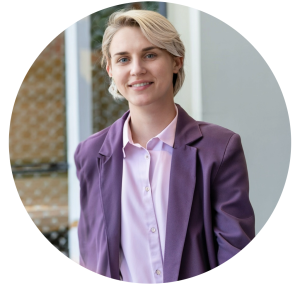 Zeke, a young adult, is wearing a purple blazer and lilac shirt and looking pleased. 